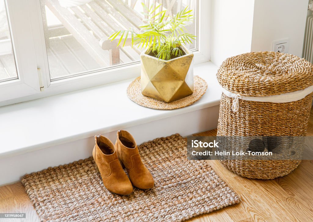 Hardwood floor with jute doormat, shoes and flower pot and seagrass laundry basket by window. Natural material objects in home concept. Home interior. Doormat Stock Photo