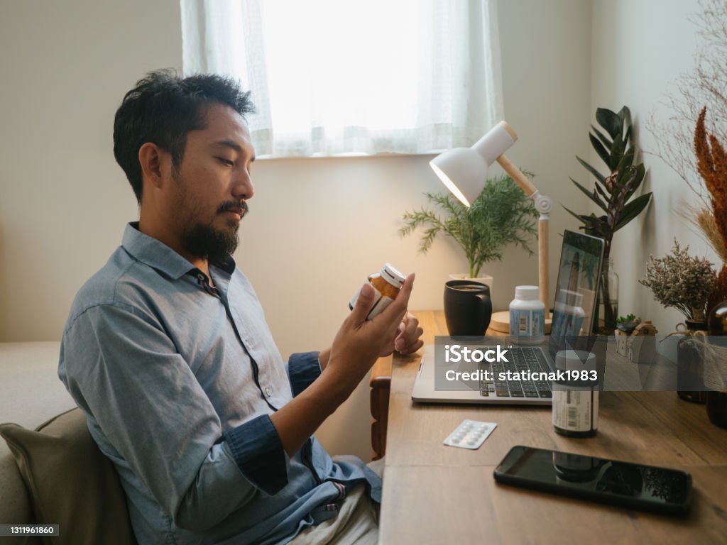 Asian man a video telemedicine call with a doctor. Asian man looking at pill bottle and listening to women doctor in consultation medicine to video conference at home. Prescription Medicine Stock Photo