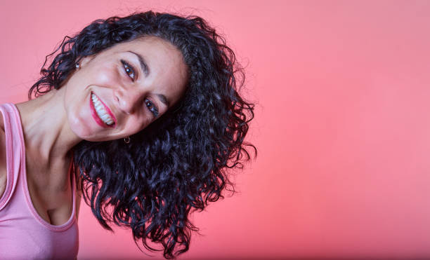 retrato de uma jovem mulher de cabelos encaracolados preto sorrindo com seus longos cabelos para o lado olhando para a câmera em um fundo rosa. método menina encaracolado. cuidados com o cabelo e conceito de beleza - mid teens hairstyle curly hair long hair - fotografias e filmes do acervo