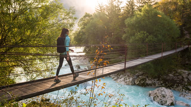 Young woman crossing a wooden suspension bridge over mountain stream, vibrant scenery