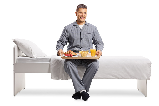 Smiling young man in pajamas sitting on a bed with breakfast on a tray isolated on white background