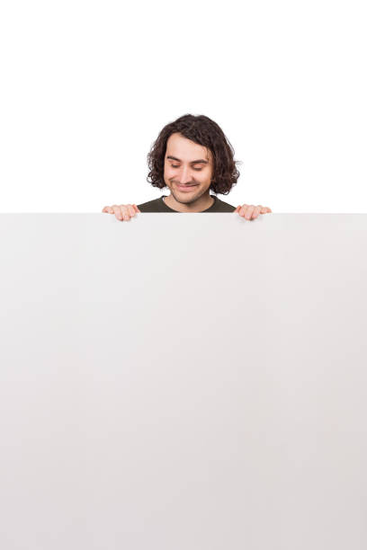 istock Positive young man, standing behind a big blank banner looking down contented at the copy space message. Cheerful male and an empty advertising placard. Happy guy reading text announcements. 1311980287