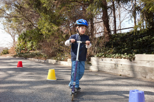 Mixed ethnic Caucasian Cuban boy (6-7 yrs old) at play riding scooter in control around cones on the move in driveway wearing safety helmet in casual clothing.
