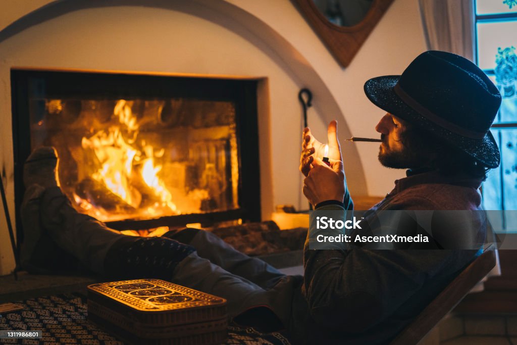 Young man relaxes by fire and smokes cbd joint He relaxes inside wintery mountain home, warming up Cannabis Plant Stock Photo