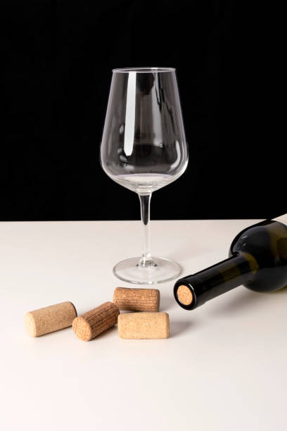 istock Bottle of white wine, wine glass and corks on white table against black background. Selective focus, minimalistic, vivid colors. 1311989764