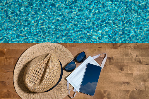 Straw hat, sunglass, smartphone and Surgical Mask on wooden floor by the pool.