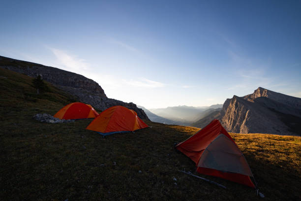 high angle view of the mountains with grass and tents in the foreground on a sunrise Morning in the mountains with three orange tents on the ground with blue sky in summer base camp stock pictures, royalty-free photos & images