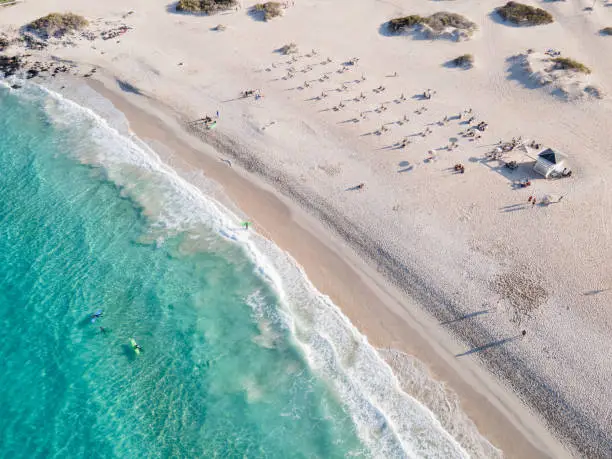 Summer image of a white sand beach with people sunbathing and surfing aerial shot. There is a lifeguard tower and the water is turquoise and clear. There are some surfers in the water. Shot in Corralejo, Fuerteventura, Canary Islands