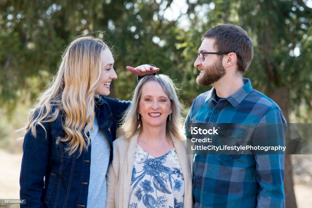 Comparing their Height to Their Mother Mother smiling a toothy smile as she is standing with her adult son and daughter for a portrait on mothers day. The young adult daughter is laughing and mimicking measuring her mother's height playfully to her brother. Short Person Stock Photo
