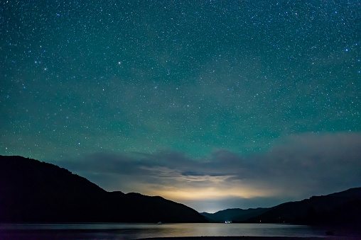 starry sky above the shape of mountains with the light of a city in a fjord in the Marlborough Sounds, New Zealand