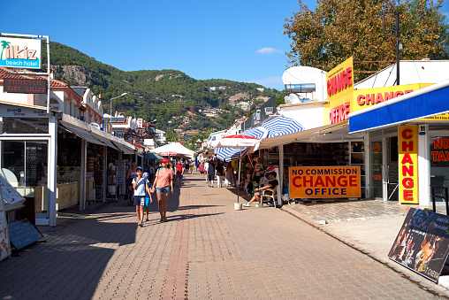 Oludeniz,  Mugla province, Turkey - Oсtober 07, 2019: People walk along the main shopping street of the resort town of Oludeniz on a hot sunny day. Sunlight and blue sky, mountains in the background