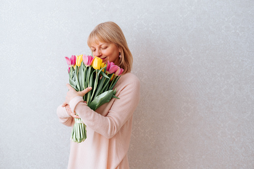 Portrait of Beautiful mature happy woman with blonde hair, smells and holding bouquet of pink and yellow tulip flowers. White background, copy space, close up.