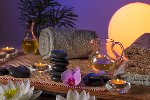 Black massage (balance) stones, Orchid flowers, towels, candles and massage oil on a wooden table with floating water lilies (lotus). Evening sun with purple sky at the background.