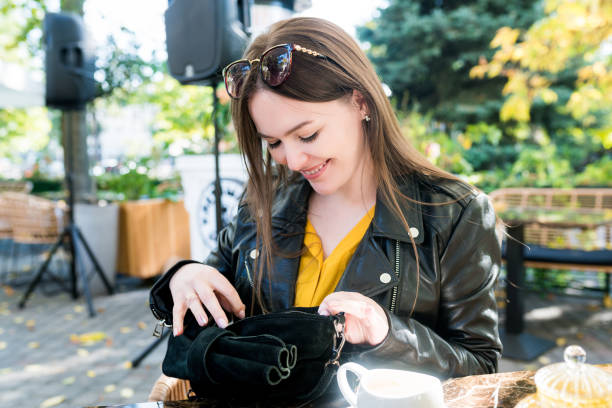 young smiling brunette woman in casual clothes sitting in street cafe and looking into the bag, seaching card to pay for lunch on sunny day - woman reaching into handbag imagens e fotografias de stock