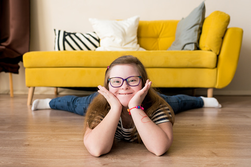 Portrait of smiling teenager girl with Down syndrome lying on the floor at home against the background of yellow sofa. Domestic life of people with disabilities.
