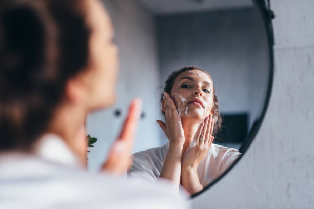 Woman washes her face in front of the mirror, applying foam to her cheeks stock photo