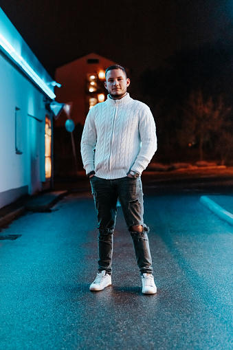 Fashionable handsome young man standing at night in front of illuminated urban gas station shop looking confident and happy towards the camera. Millennial Generation Urban Night Lifestyle Portrait.