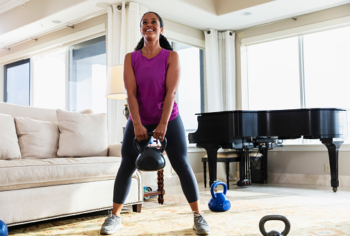 A mid adult African-American woman in her 30s exercising with kettlebells at home in the living room.