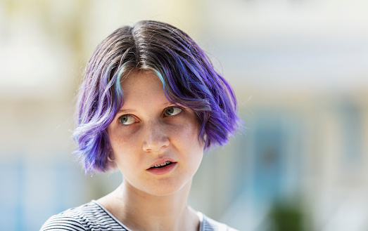 Headshot of a 13 year old teenage girl with purple hair and braces. She is standing outdoors with buildings out of focus in the background. She is rolling her eyes and frowning slightly, a typical adolescent.