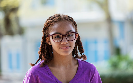 Headshot of a mixed race 13 year old tween girl wearing eyeglasses. She is standing outdoors, buildings out of focus in the background, looking at the camera.