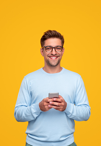 Smart young guy in glasses smiling and looking at camera while browsing mobile phone against yellow background