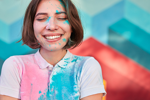Cheerful young female with closed eyes and paint on face and t shirt smiling while standing on blurred background of colorful wall
