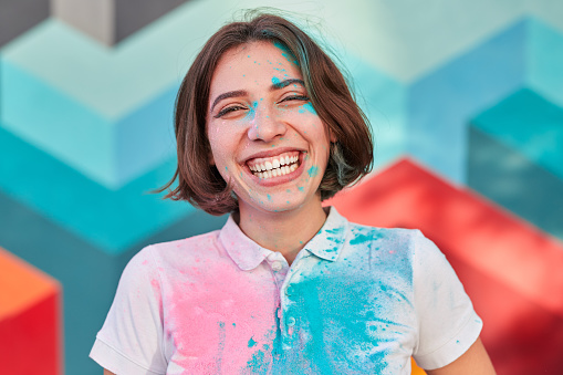 Optimistic young female with paint on face and shirt laughing happily while standing against colorful wall