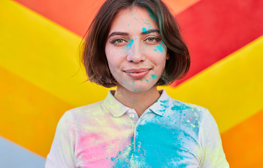 Young hipster woman covered with bright Holi festival paints looking at camera against vivid graffiti wall