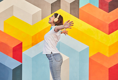 Side view of cheerful woman listening to music with headphones and spreading arms against vivid geometric graffiti