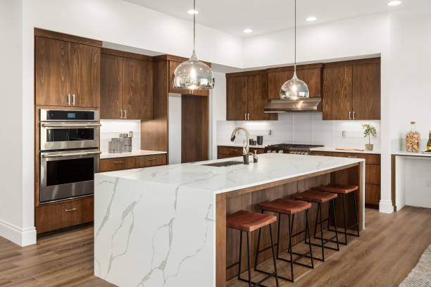 Beautiful kitchen in new luxury home with island, pendant lights, and hardwood floors. Features quartz waterfall island with dark cabinets and stainless steel appliances. stock photo