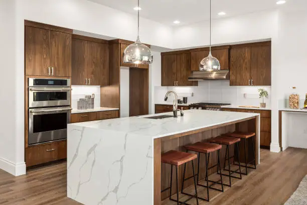 Photo of Beautiful kitchen in new luxury home with island, pendant lights, and hardwood floors. Features quartz waterfall island with dark cabinets and stainless steel appliances.