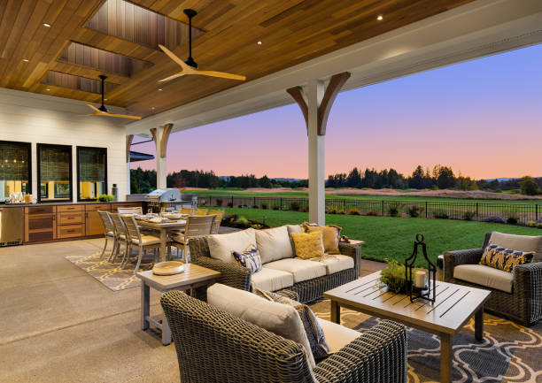 Luxury home exterior at sunset: Outdoor covered patio with kitchen, barbecue, dining table, and seating area, overlooking grass field and trees. Covered patio with beautiful sunset view porch stock pictures, royalty-free photos & images