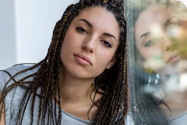 Young woman with dreadlocks and nose piercing leaning against a window with reflection on the glass looking thoughtfully at the camera in close up