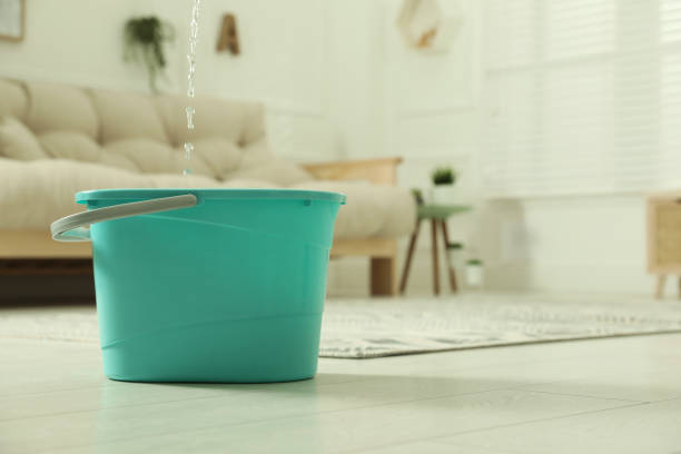 Water dripping from ceiling into plastic bucket on floor in living room, space for text. Leaking roof