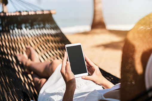 Young woman traveler lying on a hammock and using smartphone at the beach while traveling for summer vacation