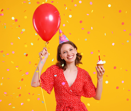 Happy young female in party hat with red balloon and cupcake having fun during birthday celebration against yellow background with confetti
