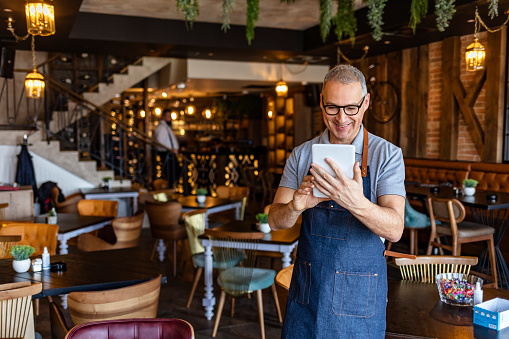 Mature waiter wearing denim apron and standing while holding digital tablet with the menu of the day.