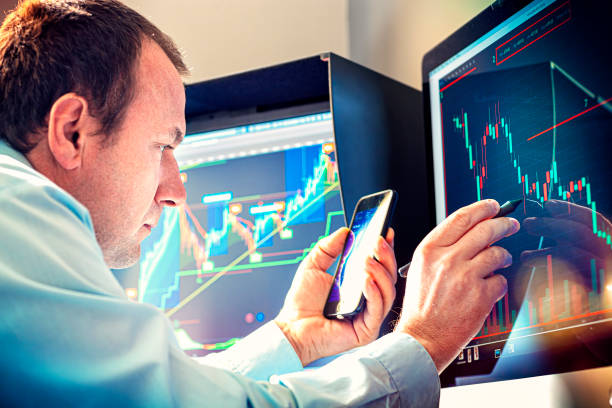 Economist with a smartphone in hand examines the economic graph in front of a computer monitor. Economist with a smartphone in hand examines the economic graph in front of a computer monitor. Stock market analyst stock trader stock pictures, royalty-free photos & images