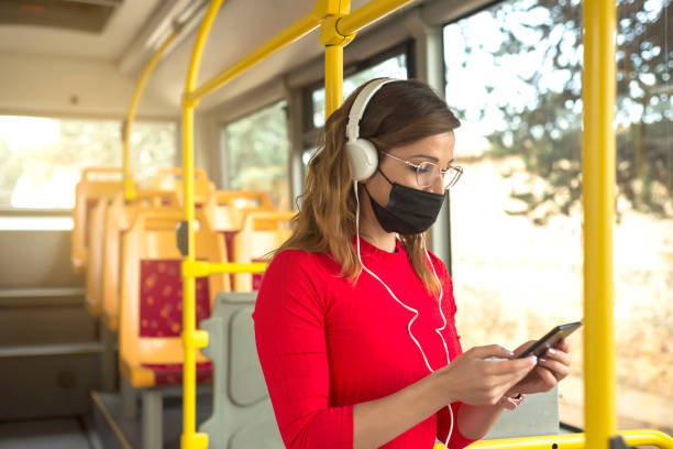 una giovane donna che indossa una maschera protettiva e cuffie bianche che ascoltano musica su un autobus pubblico. - bus transportation indoors people foto e immagini stock