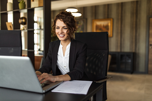 Smiling woman using laptop at work, in luxurious office, portrait.