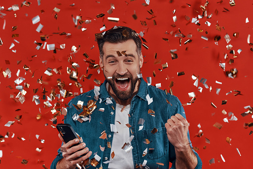 Happy young man in casual clothing holding smart phone and cheering while standing against red background with confetti flying around