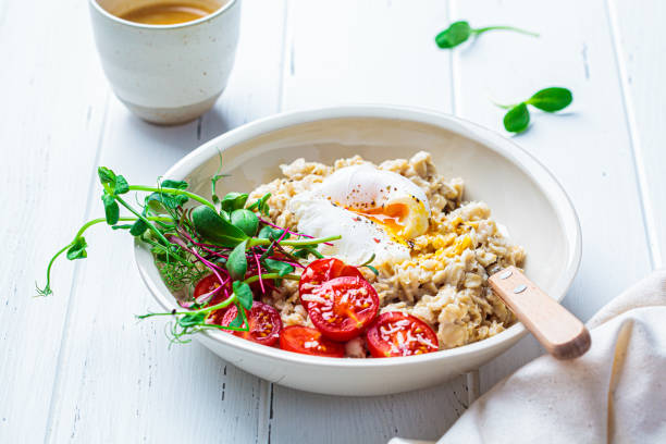 Savory oatmeal with poached egg, tomatoes, cheese and sprouts in white bowl. Healthy breakfast concept.
