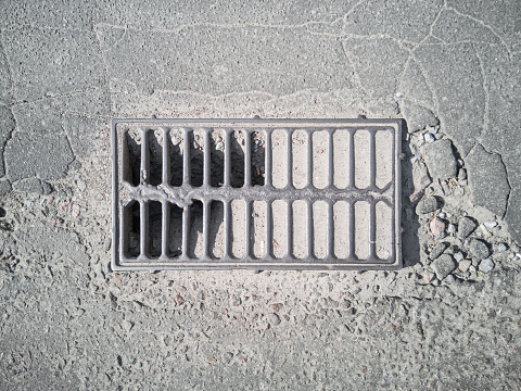 The non-functioning grate of the street stormwater storm sewer for street drainage is clogged with sand, silt and dirt on the broken asphalt, top view.