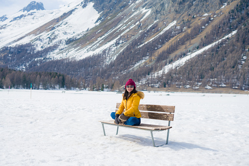 unique Winter holidays trip to amazing snow valley - young happy and beautiful Asian woman playful on bench alone enjoying snowy mountain view at Swiss Alps
