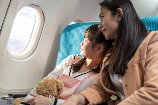 Asian mother and daughter sitting on board in airplane, looking outside window together. The girl holding bear doll looking view when the plane is flying in the sky. Airline transportation concept.