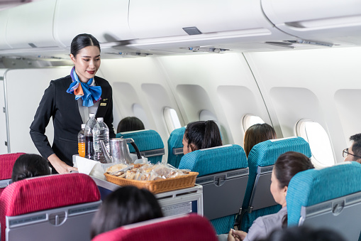 Asian female flight attendant serving food and drink to passengers on airplane. The cabin crew pushing the cart on aisle to serve the customer. Airline service job and occupation concept.