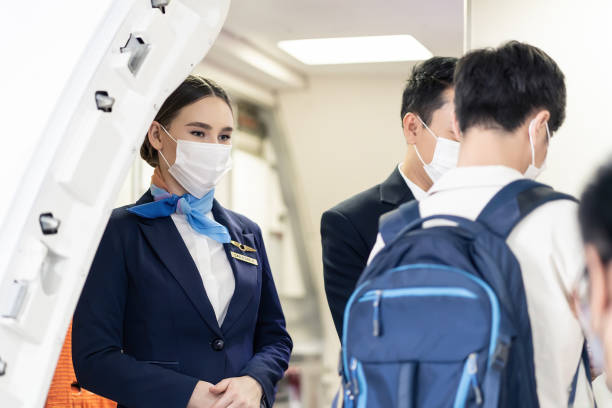 asian flight attendants wearing face mask greeting passengers walking and coming on board in airplane during the covid pandemic to prevent coronavirus infection. healthcare in transportation concept. - entering airplane imagens e fotografias de stock