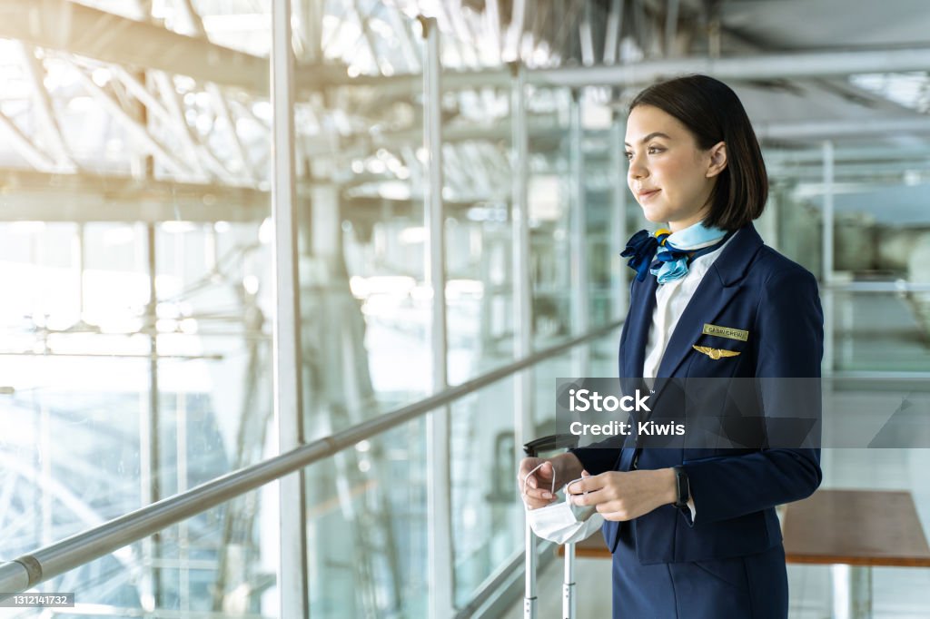 Cabin crew or air hostess holding face mask standing in passenger terminal at the airport during the COVID pandemic to prevent coronavirus infection. New normal lifestyle in air transport concept. Cabin Crew Stock Photo