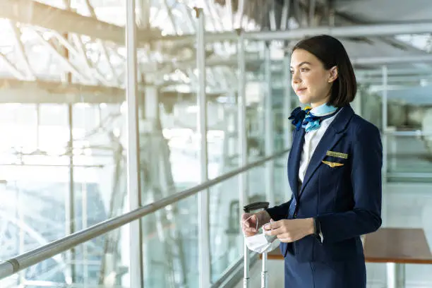 Photo of Cabin crew or air hostess holding face mask standing in passenger terminal at the airport during the COVID pandemic to prevent coronavirus infection. New normal lifestyle in air transport concept.