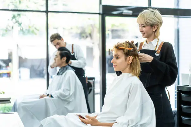 Photo of Professional young Asian female hairdresser or hair stylist talking, laughing, using scissors to cut beautiful Caucasian woman customer's blonde hair sitting with confidence in a modern beauty salon.